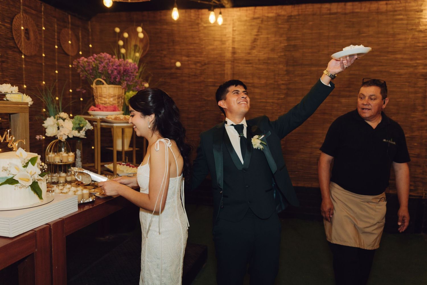 Groom playfully tossing napkin at wedding reception while bride stands near decorated cake table.