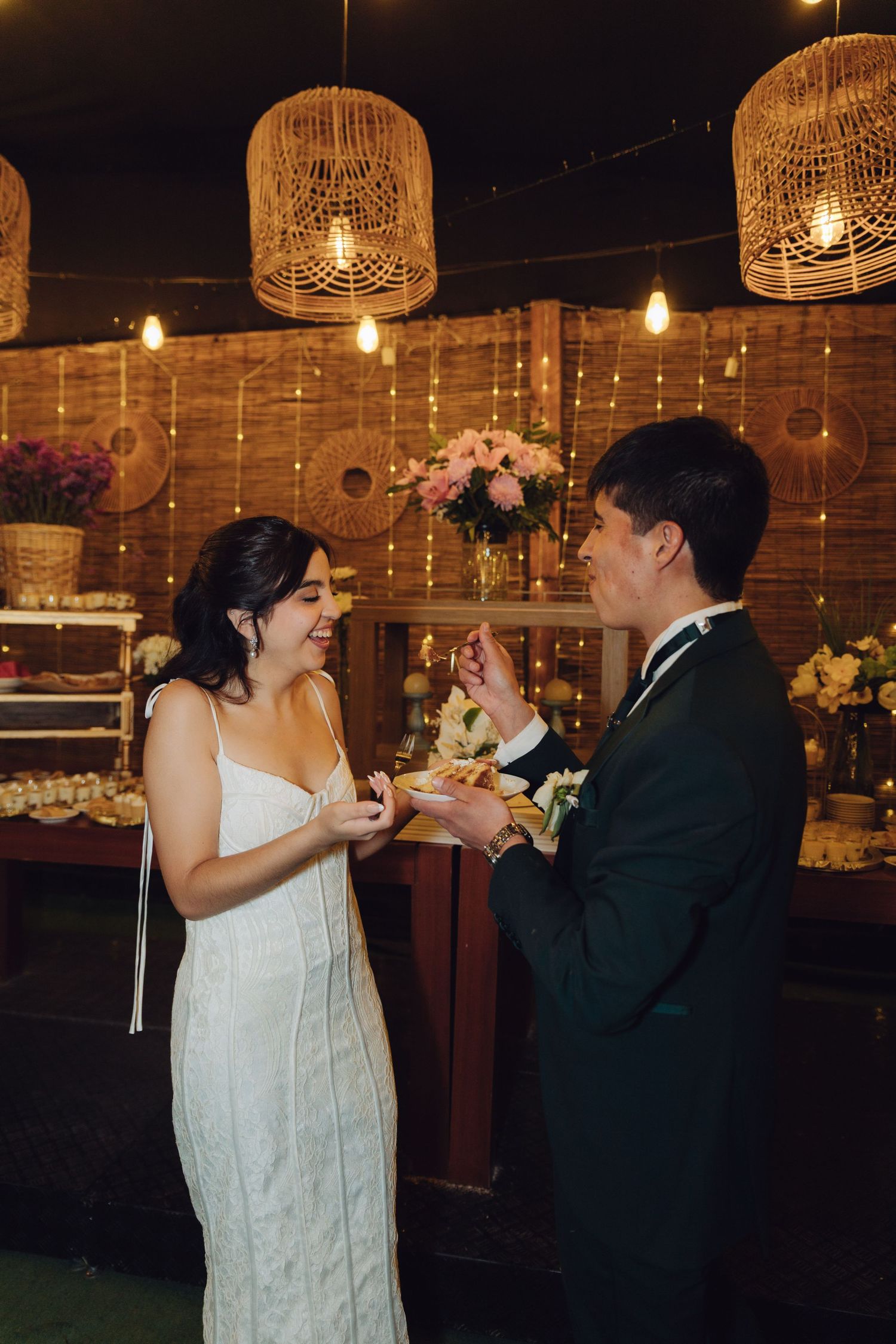 Bride and groom laughing while sharing wedding cake under warm wicker pendant lights.