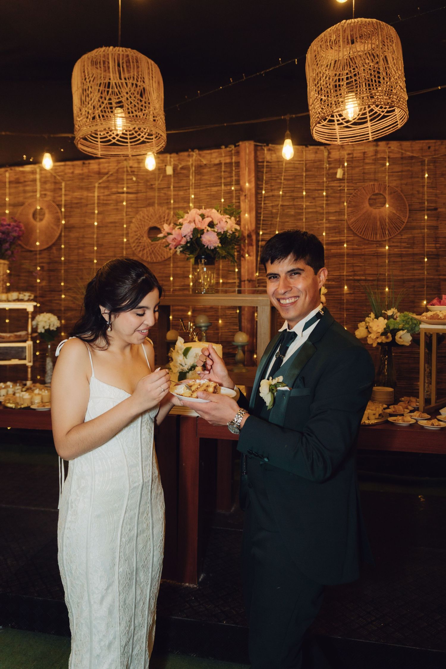 Smiling bride and groom sharing wedding cake slice under decorative wicker pendant lights.