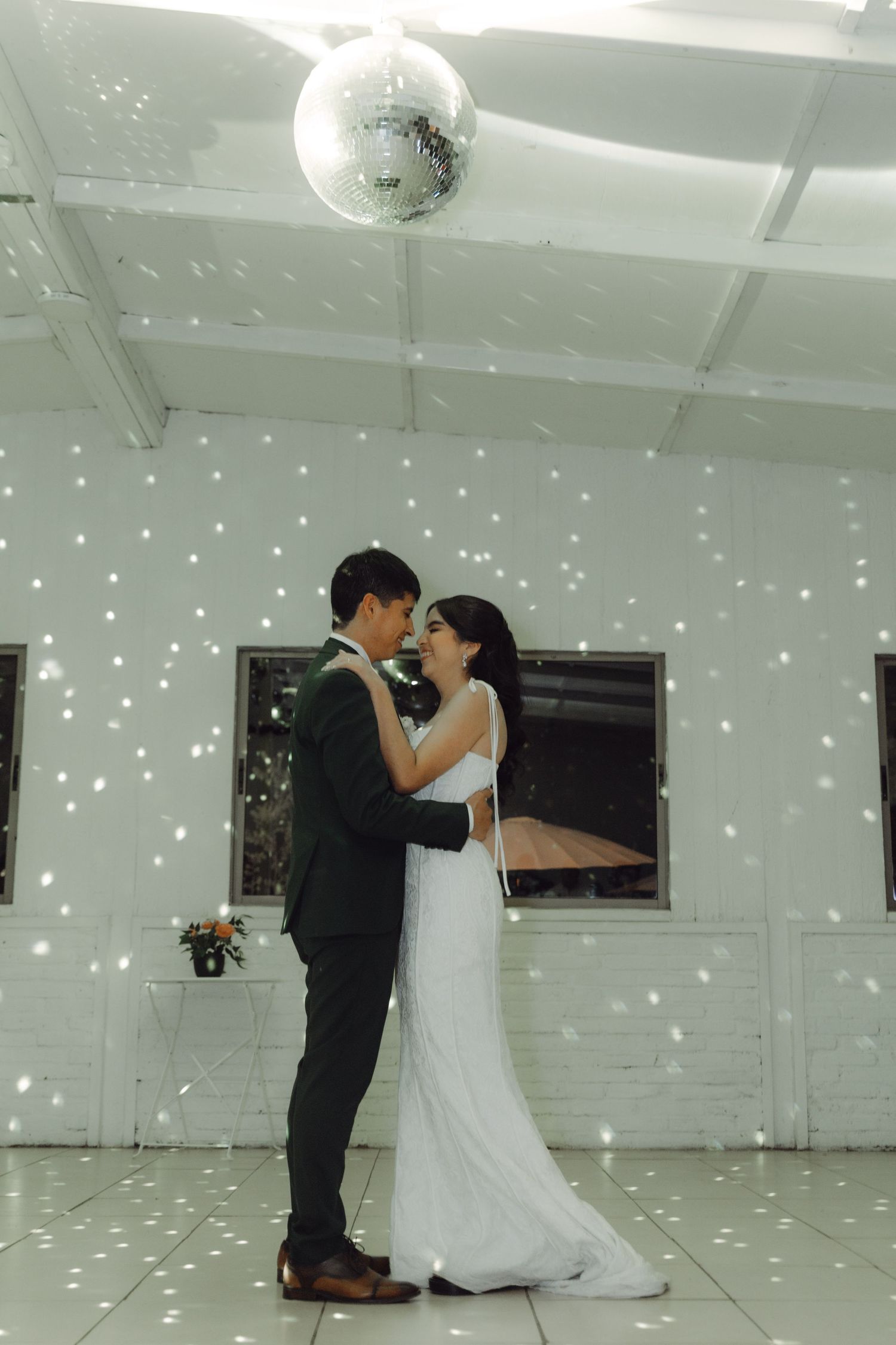 Bride and groom sharing their first dance under a sparkling disco ball in a white decorated venue.