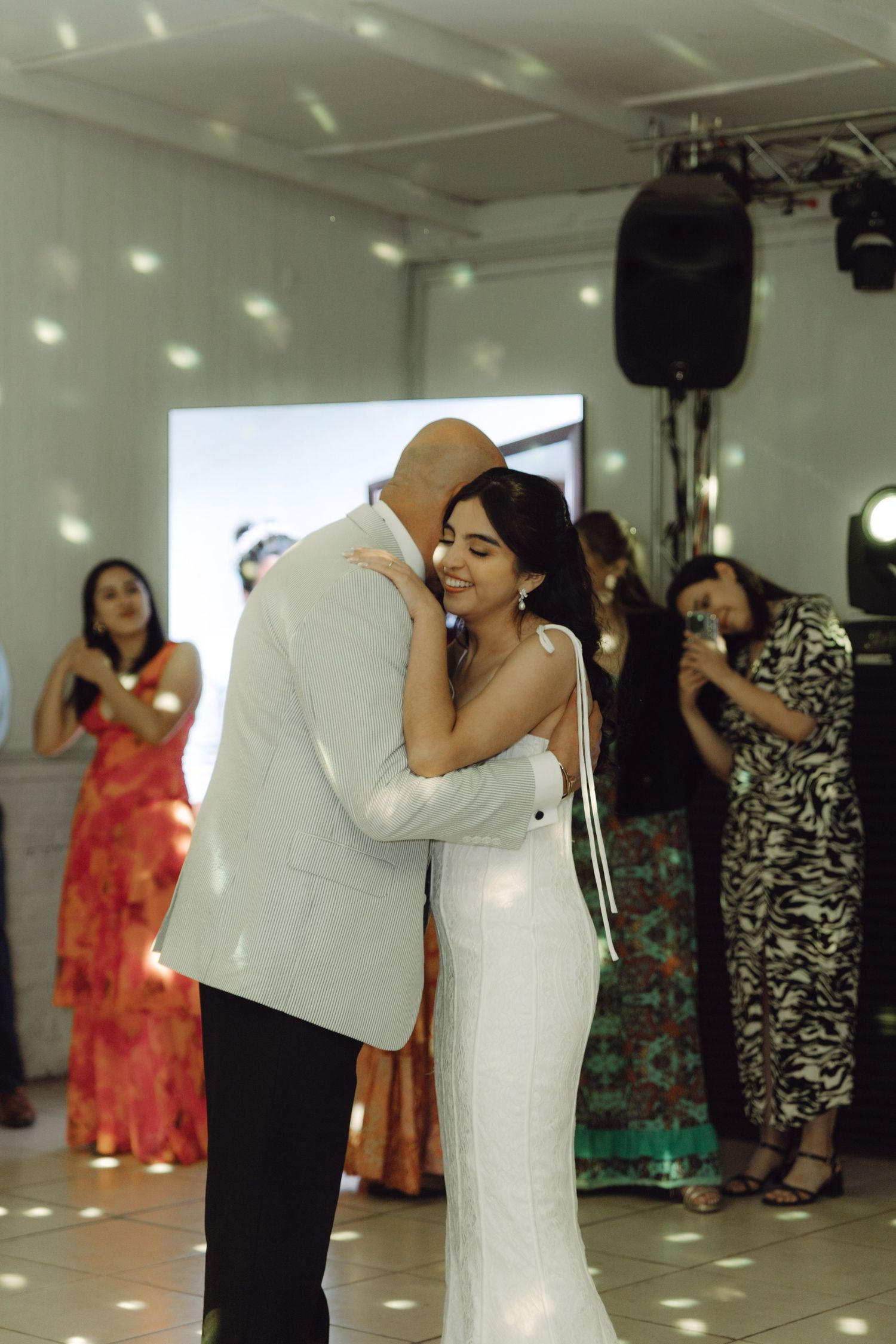Bride in white gown smiling joyfully while dancing with groom at wedding reception with guests nearby.