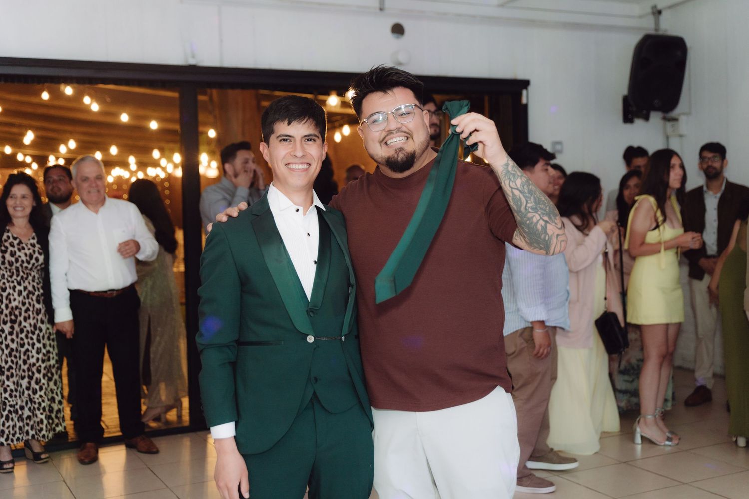 Two men posing together at a wedding reception, one holding a green tie.