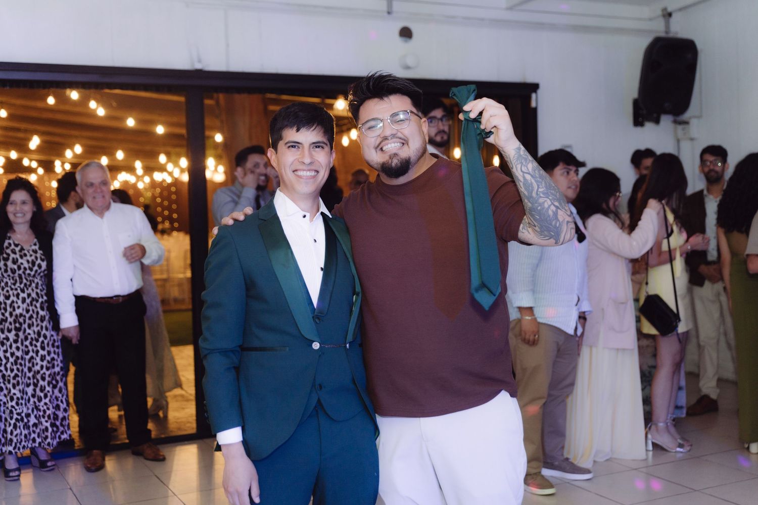 Two men smiling at a wedding reception, one holding up a green tie as a souvenir.