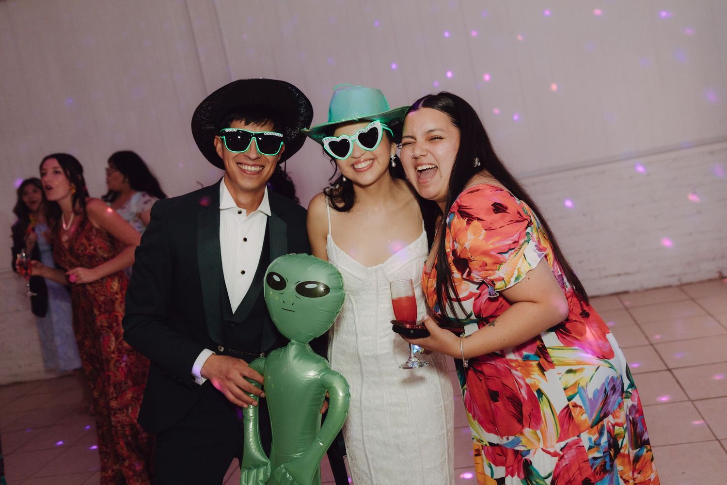 Bride, groom, and guest posing with an inflatable alien toy and fun sunglasses at wedding.