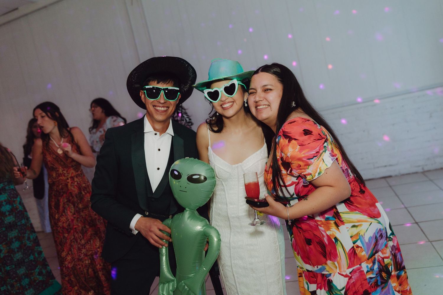 Bride and groom wearing fun sunglasses pose with inflatable alien at wedding reception dance floor.