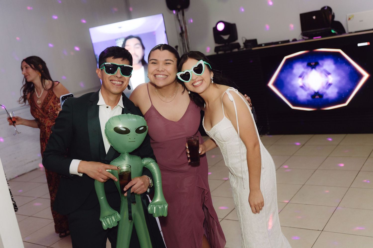 Groom and guest in sunglasses pose with inflatable alien beside smiling bride at wedding.