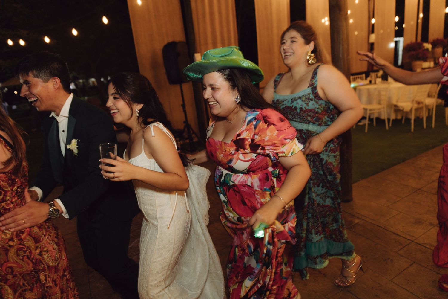 Guests dancing joyfully at a wedding reception, including a bride in white and a woman in a colorful floral dress with green hat.