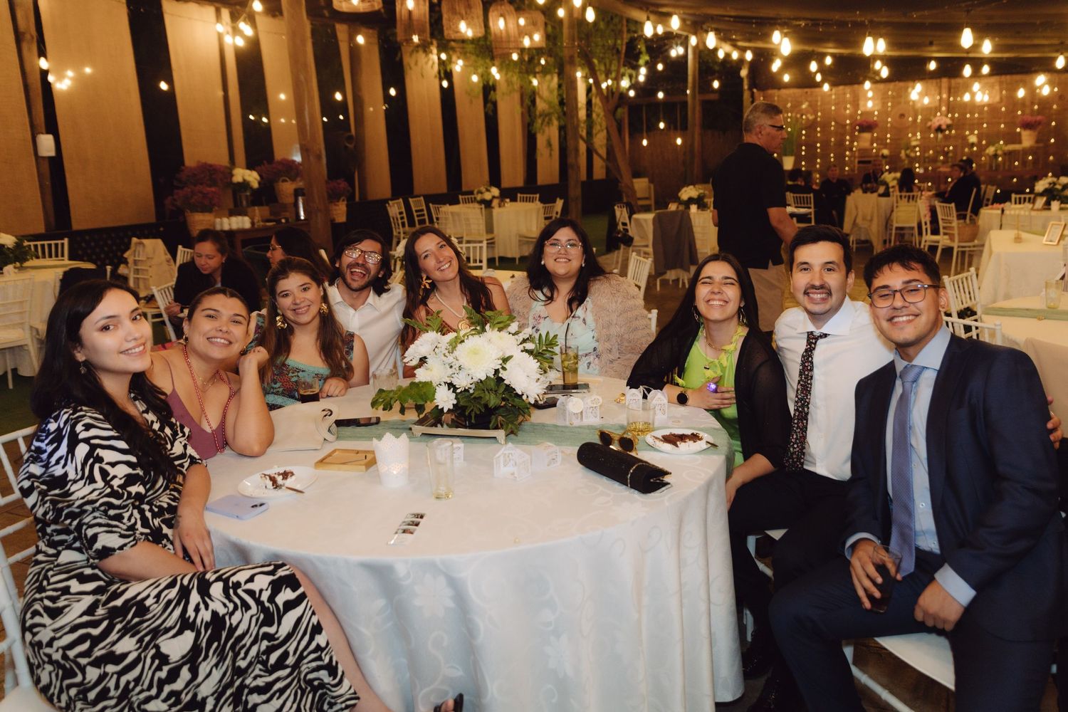 Group of elegantly dressed wedding guests smiling together at a round table decorated with white flowers and string lights.
