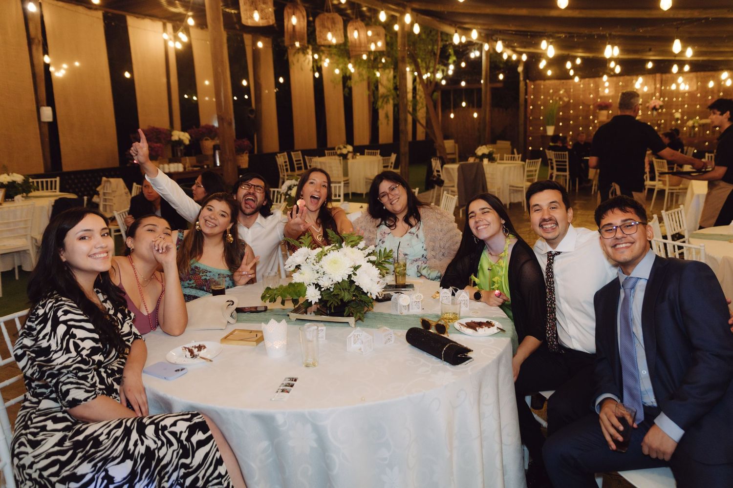 Happy group of wedding guests posing enthusiastically at a decorated reception table with floral centerpiece and warm string lights.