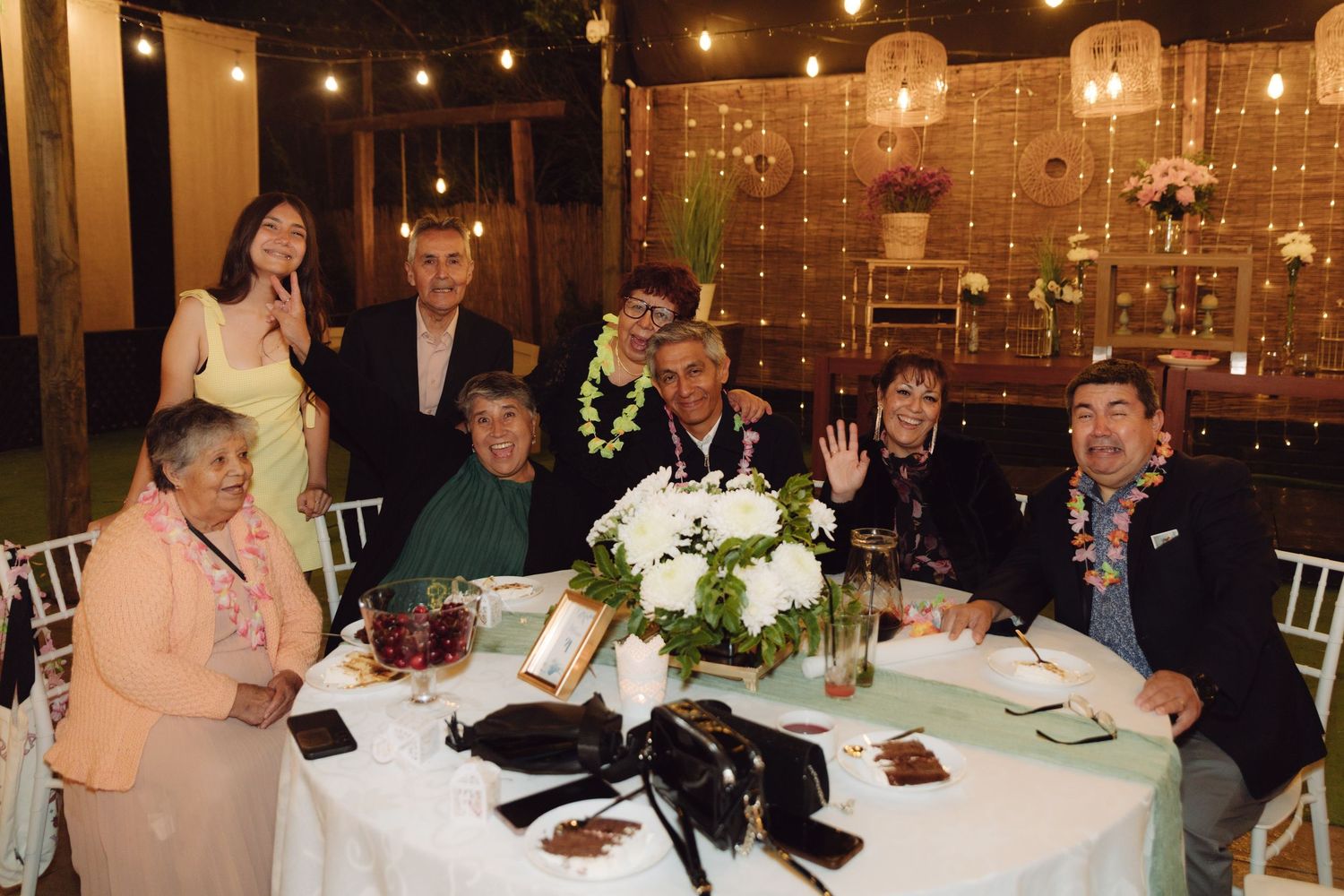 Multi-generational family group posing together at a wedding reception table with floral centerpiece and decorative lighting.