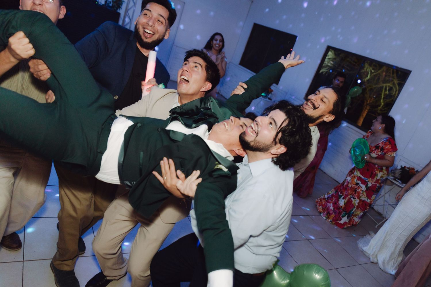 Excited wedding guests lifting a smiling man in a green suit on the dance floor surrounded by balloons.