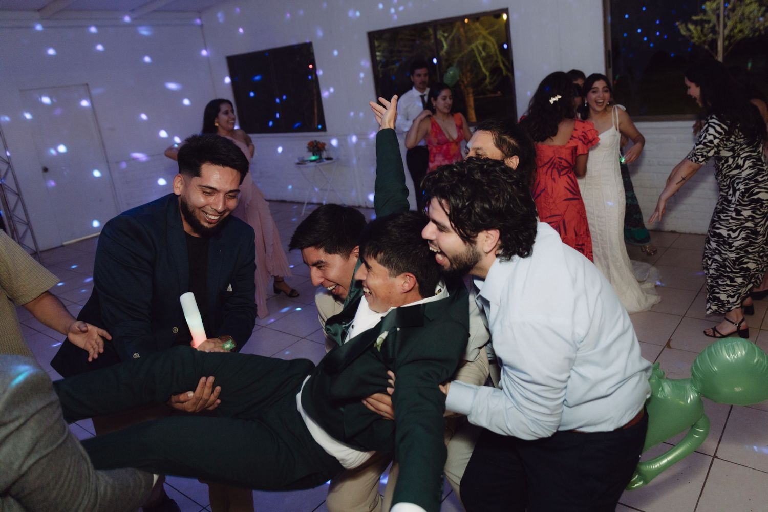 Wedding guests playfully lifting a young man in a school uniform on the dance floor during a lively celebration.
