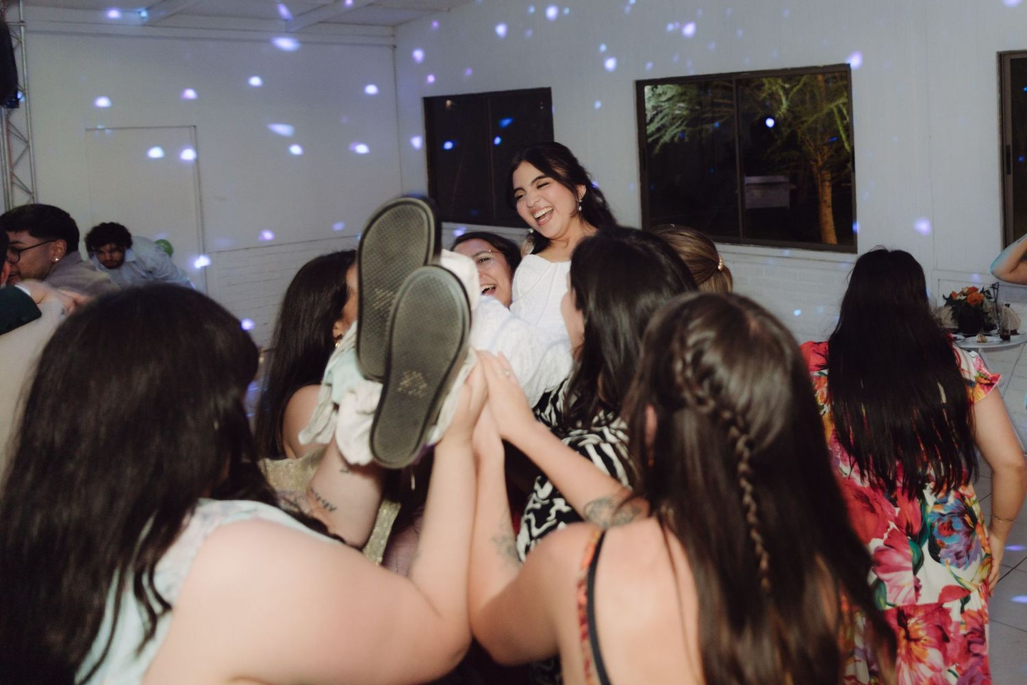 Smiling bride in white dress being lifted by friends on the dance floor during a festive wedding reception.