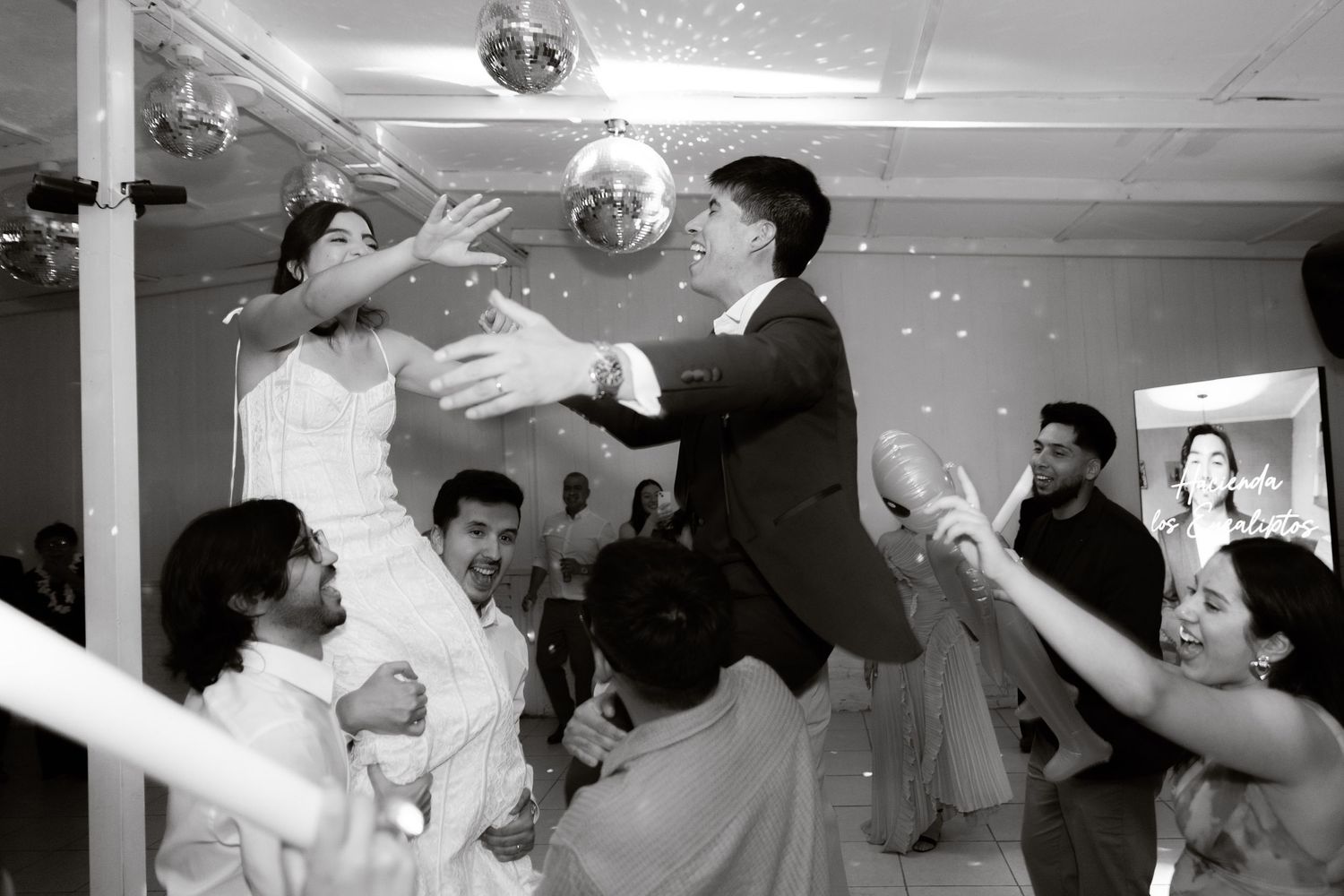 Black and white photo of bride and groom being lifted by wedding guests during an energetic reception celebration.