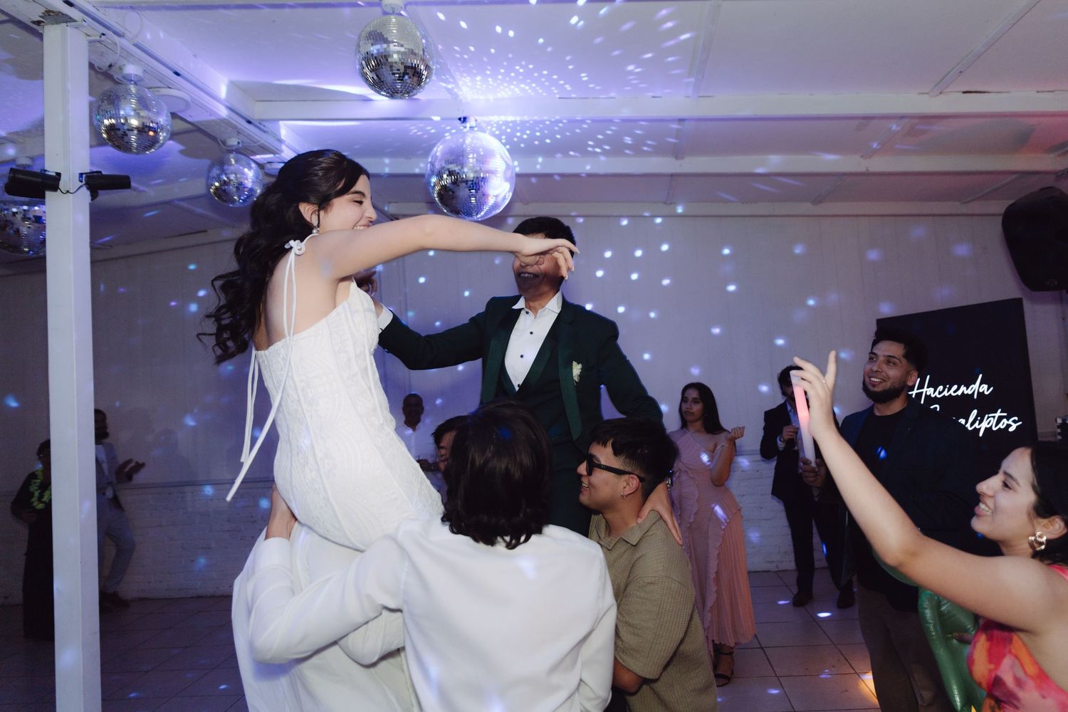 Bride lifted by guests on wedding dance floor under disco ball lights.