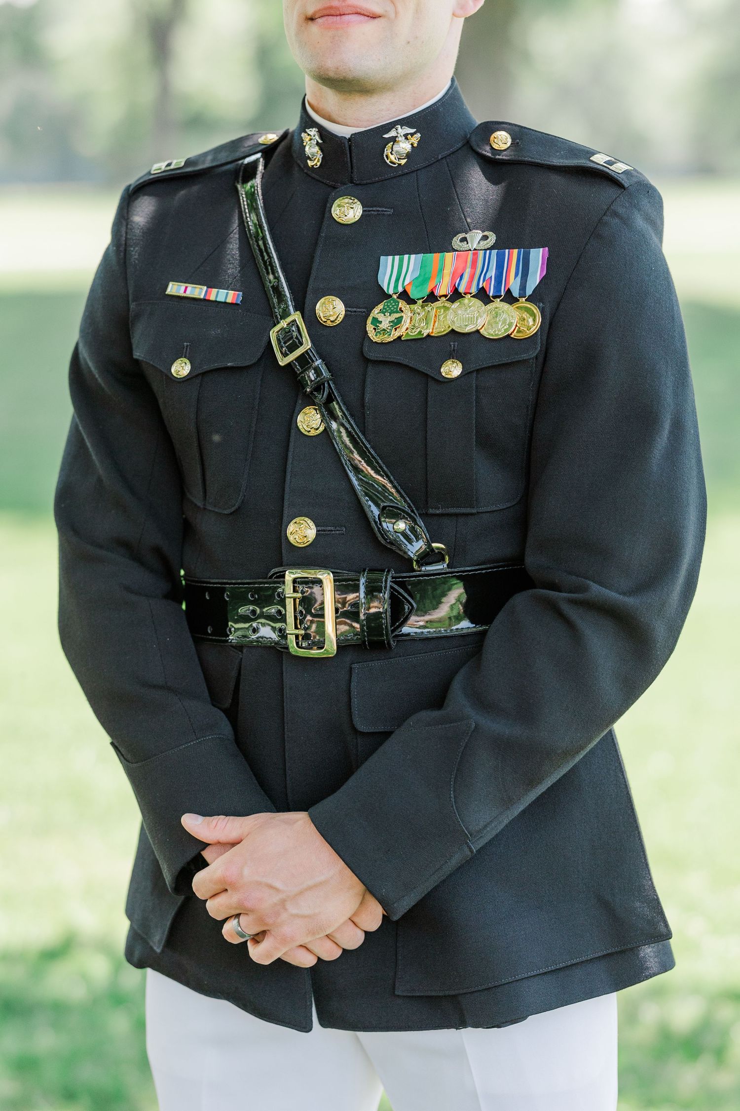 Marine officer in dress blues uniform with medals, ribbons, and gold buttons stands at parade rest outdoors.