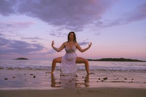 Person in white dress poses playfully on beach at sunset with dramatic purple sky.