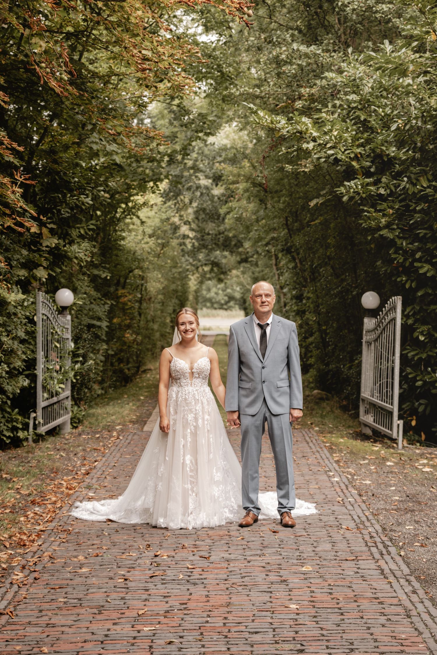 Braut im Spitzenkleid steht mit ihrem Vater im grauen Anzug auf einem Backsteinweg zwischen geöffneten Gartentoren.
