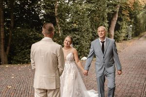 Brautvater im grauen Anzug führt seine Tochter im weißen Spitzenkleid auf einem von Bäumen gesäumten Weg zum First Look mit dem Bräutigam.
