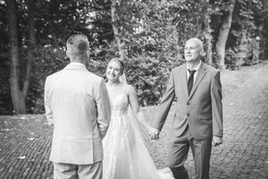 Schwarz-Weiß-Foto des Brautvaters, der seine lächelnde Tochter im Brautkleid auf einem von Bäumen gesäumten Weg zum Bräutigam begleitet.