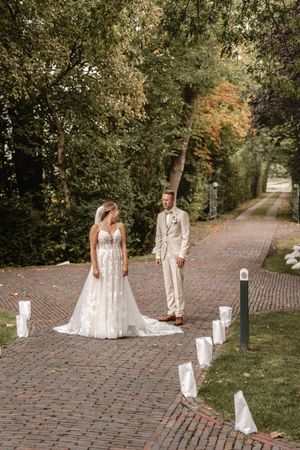 Braut im weißen Spitzenkleid und Bräutigam im beigen Anzug stehen auf einem Backsteinweg, der von Papierlaternen gesäumt ist.