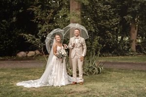 Lächelndes Brautpaar im beigen Anzug und weißen Brautkleid steht nach der Hochzeit mit durchsichtigen Regenschirmen in einem grünen Garten.