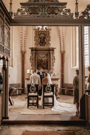 Brautpaar sitzt während der kirchlichen Trauung auf festlich geschmückten Stühlen vor einem kunstvoll verzierten Kirchenaltar.