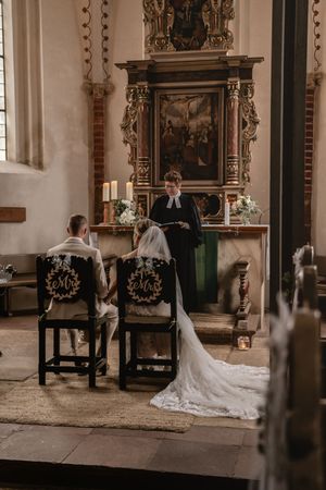Brautpaar sitzt auf festlich geschmückten Stühlen vor dem Geistlichen in schwarzer Robe am reich verzierten Kirchenaltar. Kirche Esenshamm
