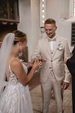 Braut im Spitzenkleid und Schleier steckt ihrem lächelnden Bräutigam während der kirchlichen Trauung den Ehering an. St. Matthäus Kirche
