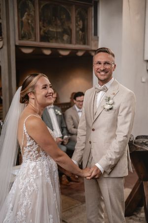 Braut und Bräutigam lächeln sich an und halten sich während der kirchlichen Trauung vor dem Altar an den Händen, während die Gäste zuschauen