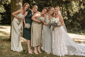 Braut im Spitzenkleid und Schleier lacht mit vier Brautjungfern in salbeigrünen Kleidern, die ihre Blumensträuße unter Bäumen halten.