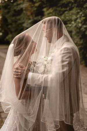 Braut und Bräutigam stehen sich unter einem transparenten Hochzeitsschleier gegenüber, umgeben von weichem Licht und grünem Blattwerk. Diele