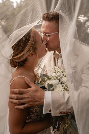 Braut und Bräutigam küssen sich unter einem transparenten Hochzeitsschleier und halten einen weißen Brautstrauß im warmen Tageslicht.