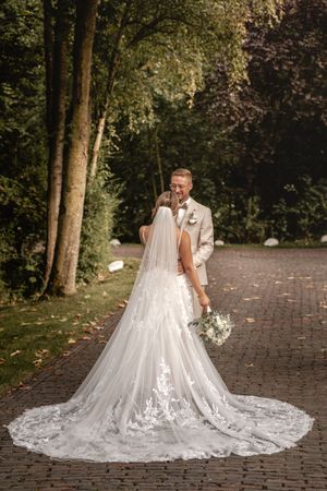 Braut im weißen Spitzenkleid mit langer Schleppe umarmt ihren Bräutigam auf einem Backsteinweg zwischen hohen Bäumen. Mittenfelder Diele
