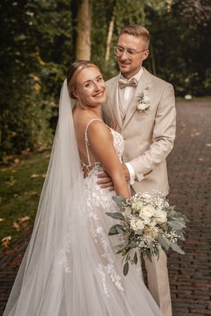 Lächelnde Braut im Spitzenkleid mit langem Schleier steht mit ihrem Bräutigam im beigen Anzug auf einem von Bäumen gesäumten Weg. Stadland