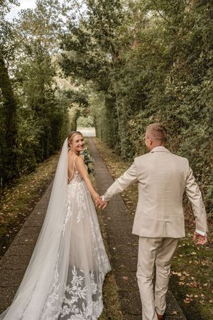 Braut im weißen Spitzenkleid mit langem Schleier blickt lächelnd zurück, während sie Hand in Hand mit ihrem Bräutigam einen Weg entlanggeht.