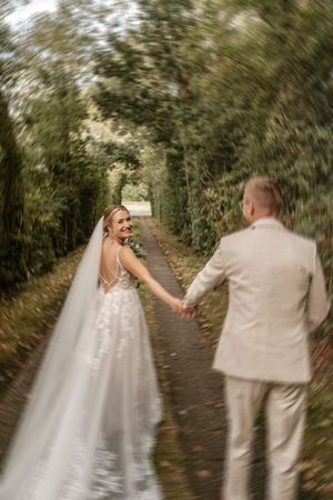 Bewegungsunschärfe in einer emotionalen Hochzeitsaufnahme: Die Braut mit langem Schleier führt ihren Bräutigam einen Waldweg entlang.