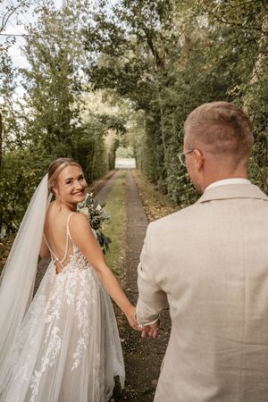 Lächelnde Braut im weißen Brautkleid und Schleier hält ihren Brautstrauß und die Hand ihres Bräutigams auf einem von Bäumen gesäumten Weg.