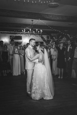 Schwarz-Weiß-Fotografie des Brautpaares beim ersten Hochzeitstanz unter Lichterketten, während die Gäste zuschauen. Mittenfelder Diele