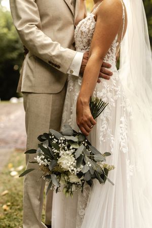 Braut im weißen Spitzenkleid hält einen Strauß aus Eukalyptus und weißen Rosen, während der Bräutigam im beigen Anzug sie umarmt. Stadland