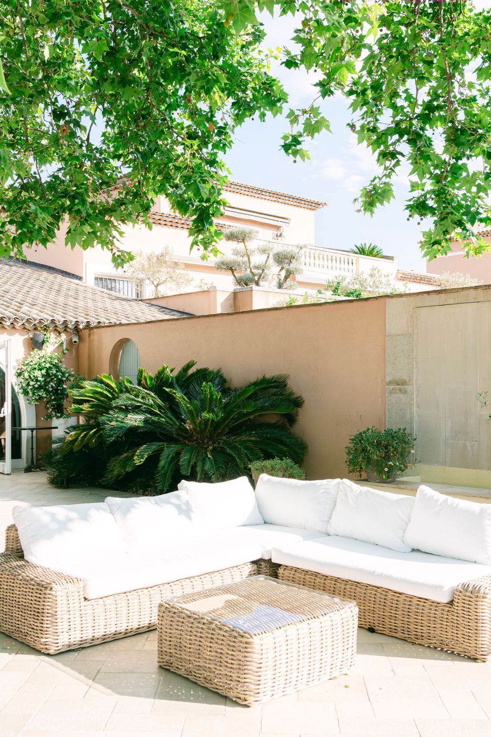 Elegant outdoor patio with wicker sectional sofa, white cushions, and coffee table surrounded by lush greenery.