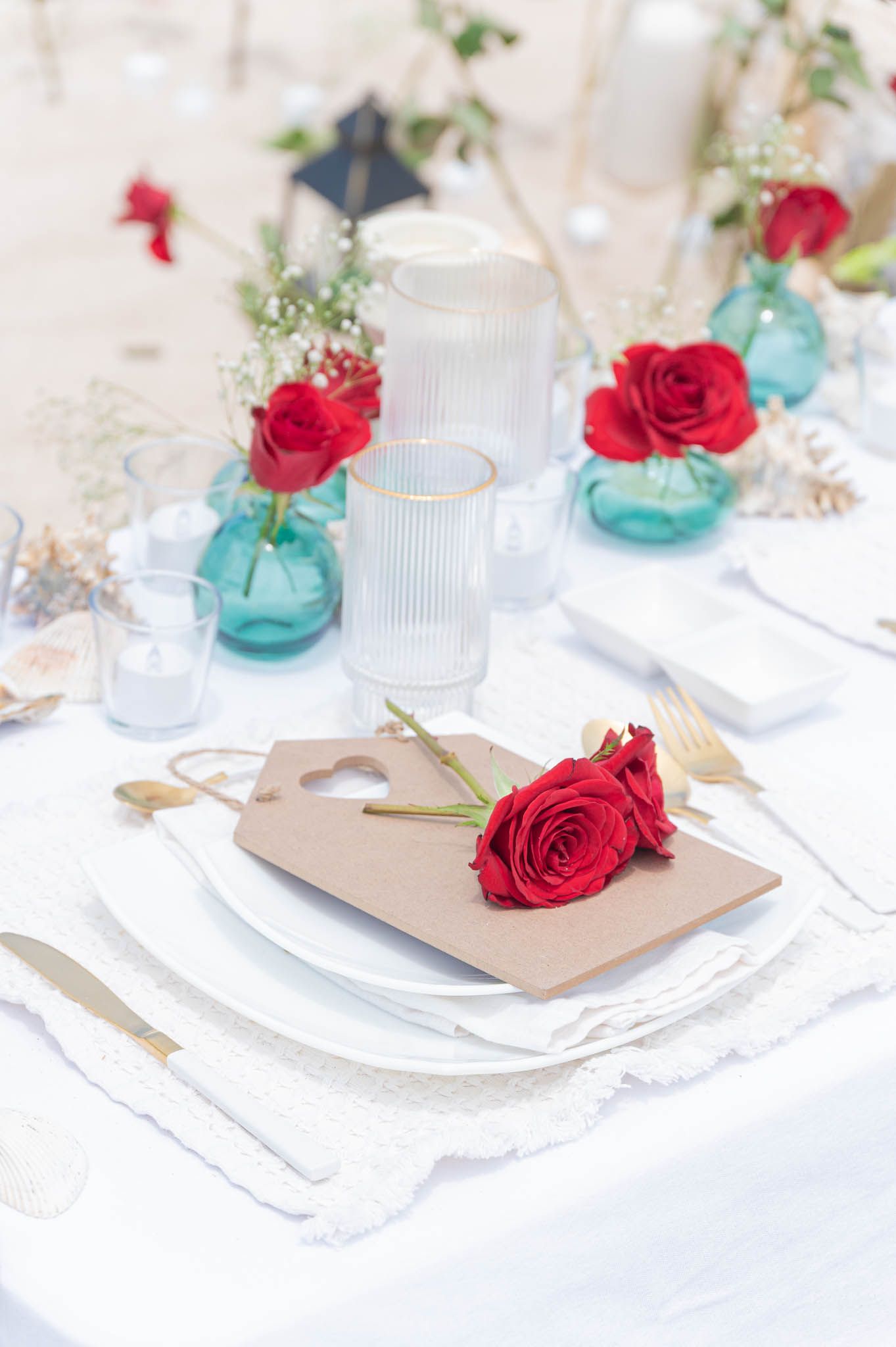 Elegant wedding table setting with red roses, teal vases, gold cutlery, and a kraft paper place card on white linens.