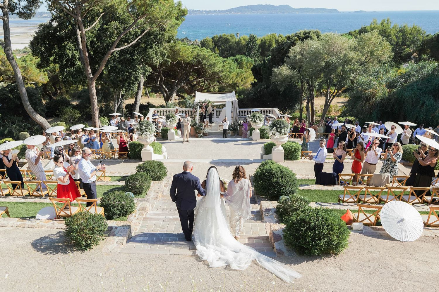 Aerial view of an elegant outdoor wedding ceremony on a terrace overlooking the sea, with guests holding parasols.