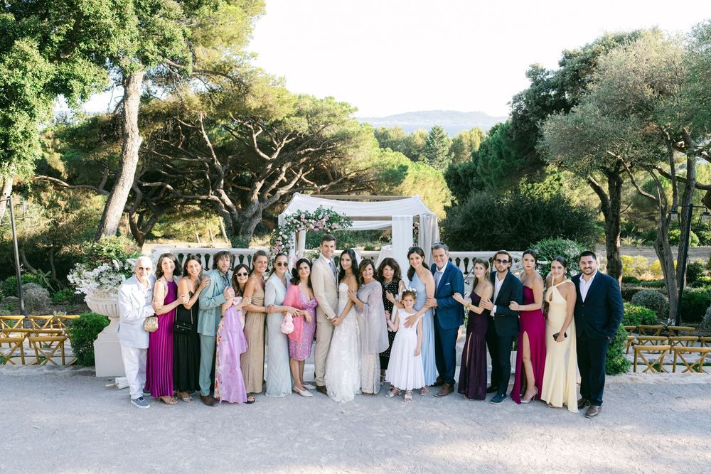 Wedding party poses outdoors at elegant venue with white arch, colorful bridesmaid dresses, and lush greenery in background.