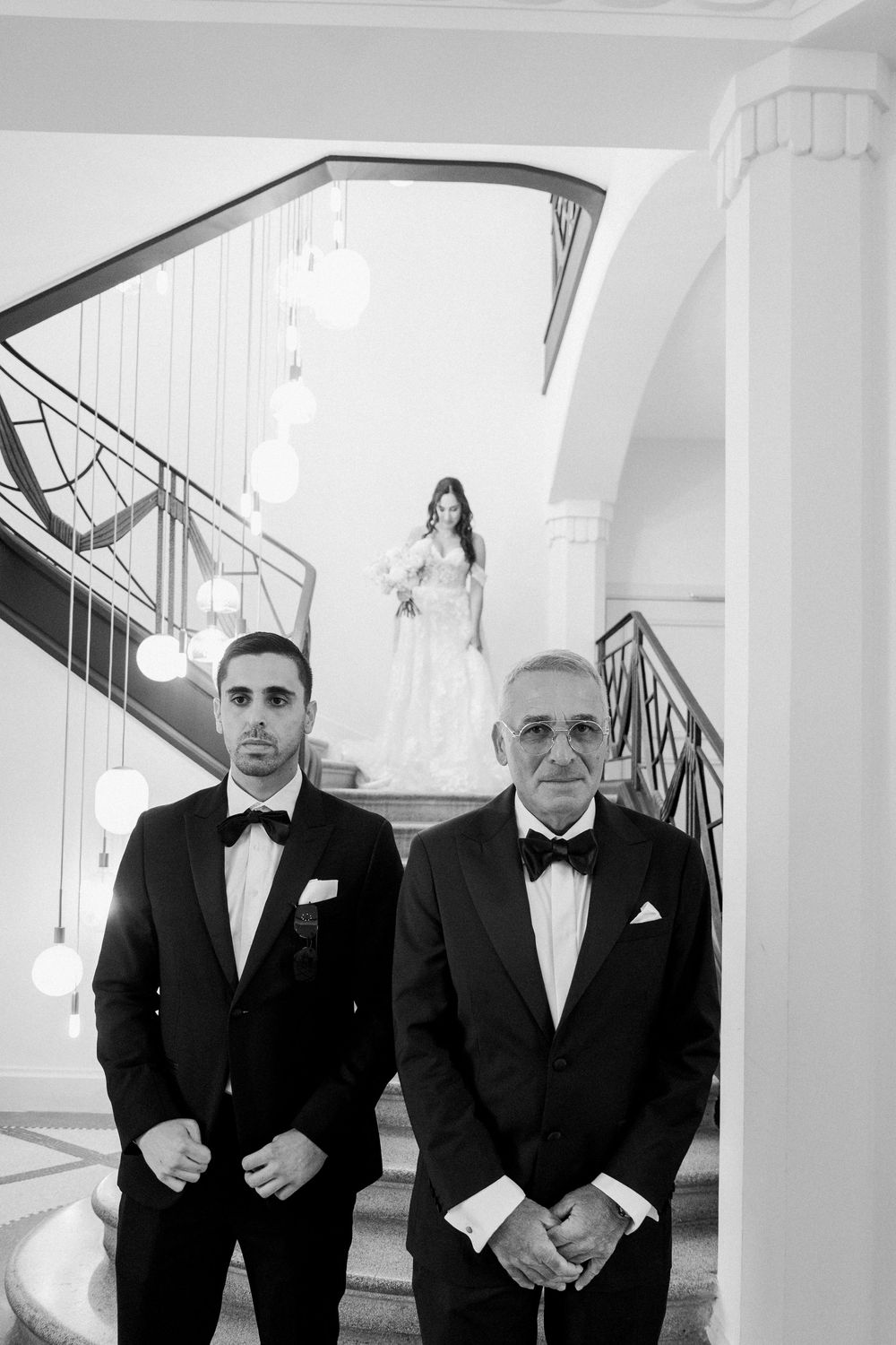 Black and white wedding photo of groom and older man in tuxedos standing before bride descending elegant staircase.