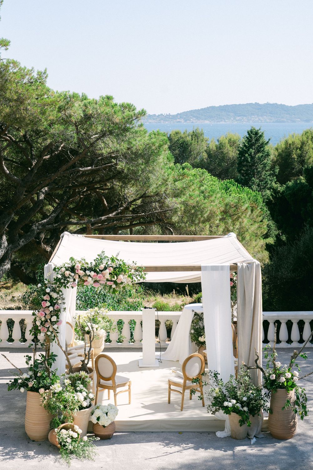 Elegant outdoor wedding ceremony setup with white draping, floral arch, gold chairs, and lush greenery overlooking a scenic landscape.