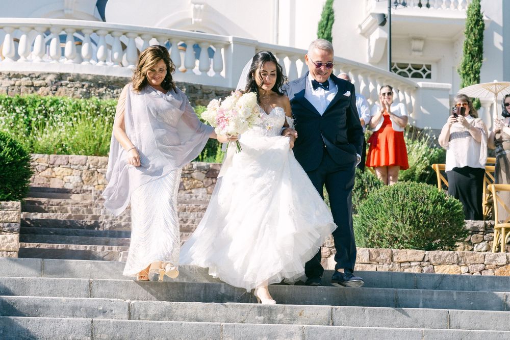 Bride in white gown walks down stone steps with parents at elegant outdoor wedding venue.