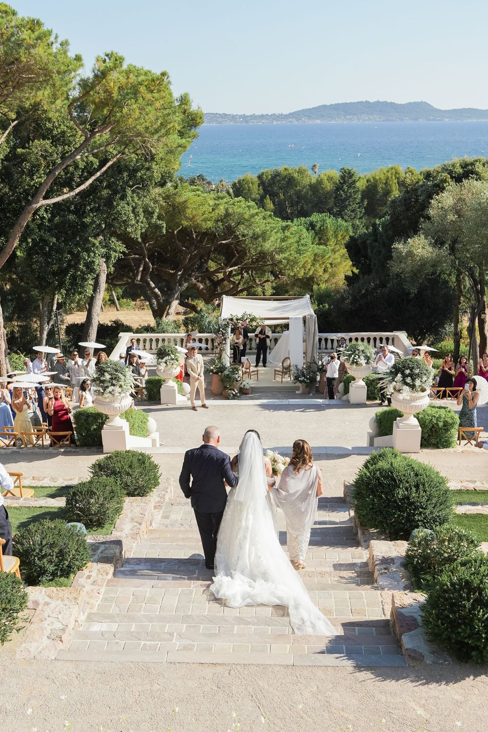 Bride walks down outdoor aisle with parents toward floral arch, overlooking Mediterranean sea at elegant garden wedding.