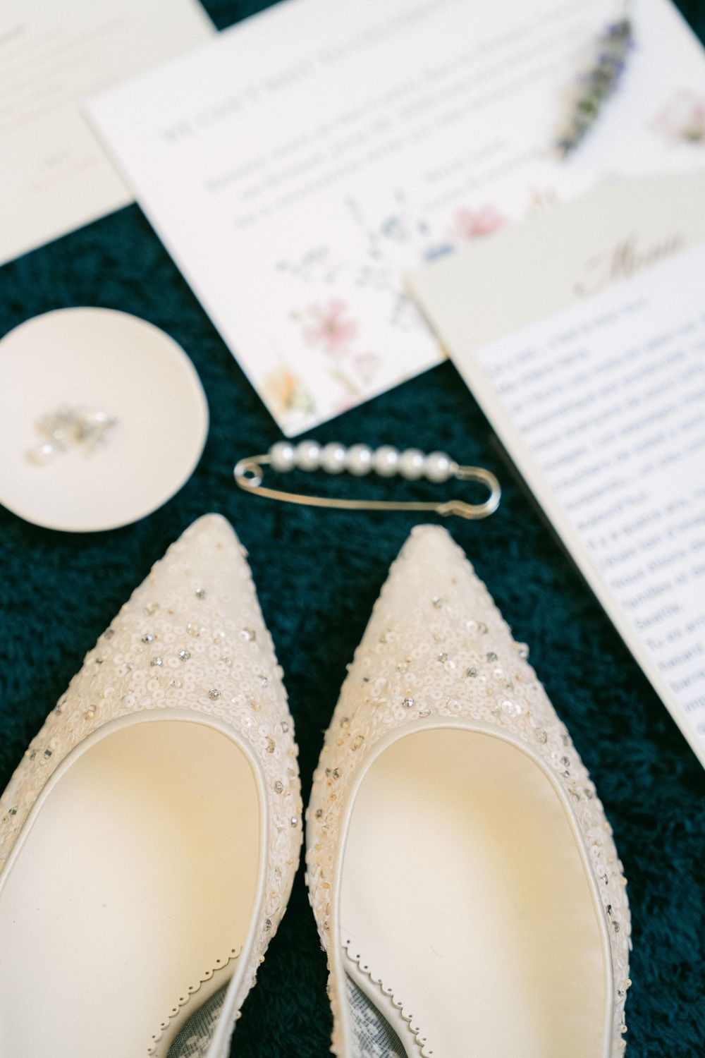 Elegant white beaded bridal heels displayed with pearl brooch, wedding invitation, and rings on dark teal surface.
