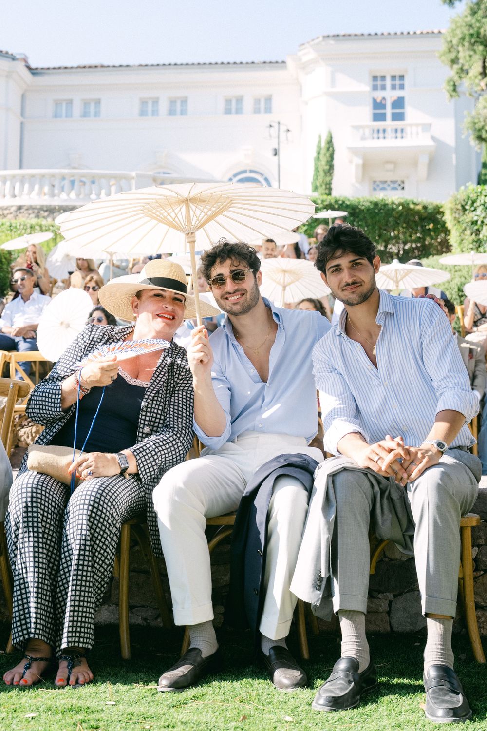 Three guests smile at an outdoor wedding reception, seated on chairs with parasols and a white villa in the background.